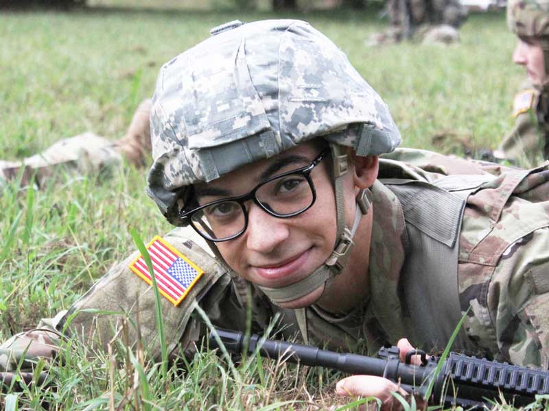 a smiling student cadet lying in the grass carrying a gun during a drill
